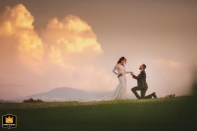 Groom kneeling and holding the bride’s hand for a couple’s portrait at Castello di Abbadia Ombrone in Tuscany, with a dramatic sky in the background.